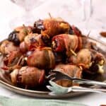 A plate of baked bacon-wrapped stuffed dates with tongs for serving.