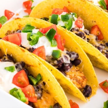 Baked chicken tacos in a baking dish topped with sour cream, chopped tomatoes, and sliced jalapenos.