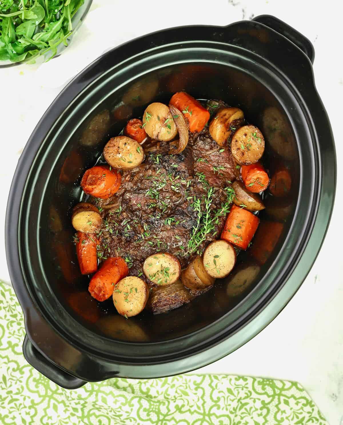 A crock-pot with a pot roast surrounded by potatoes and carrots.