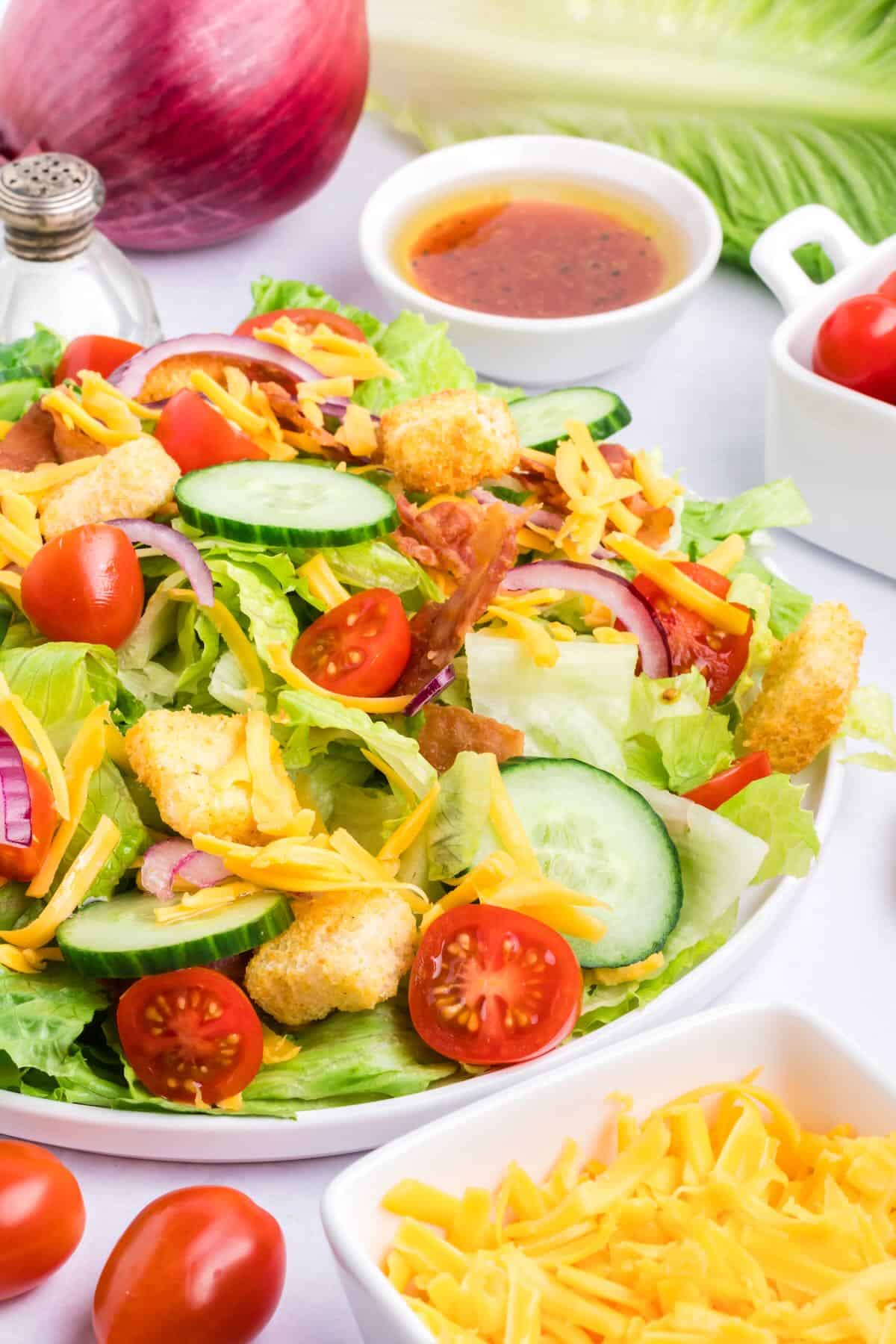 A house salad on a white plate with a bowl of dressing behind it and shredded cheese in front. 