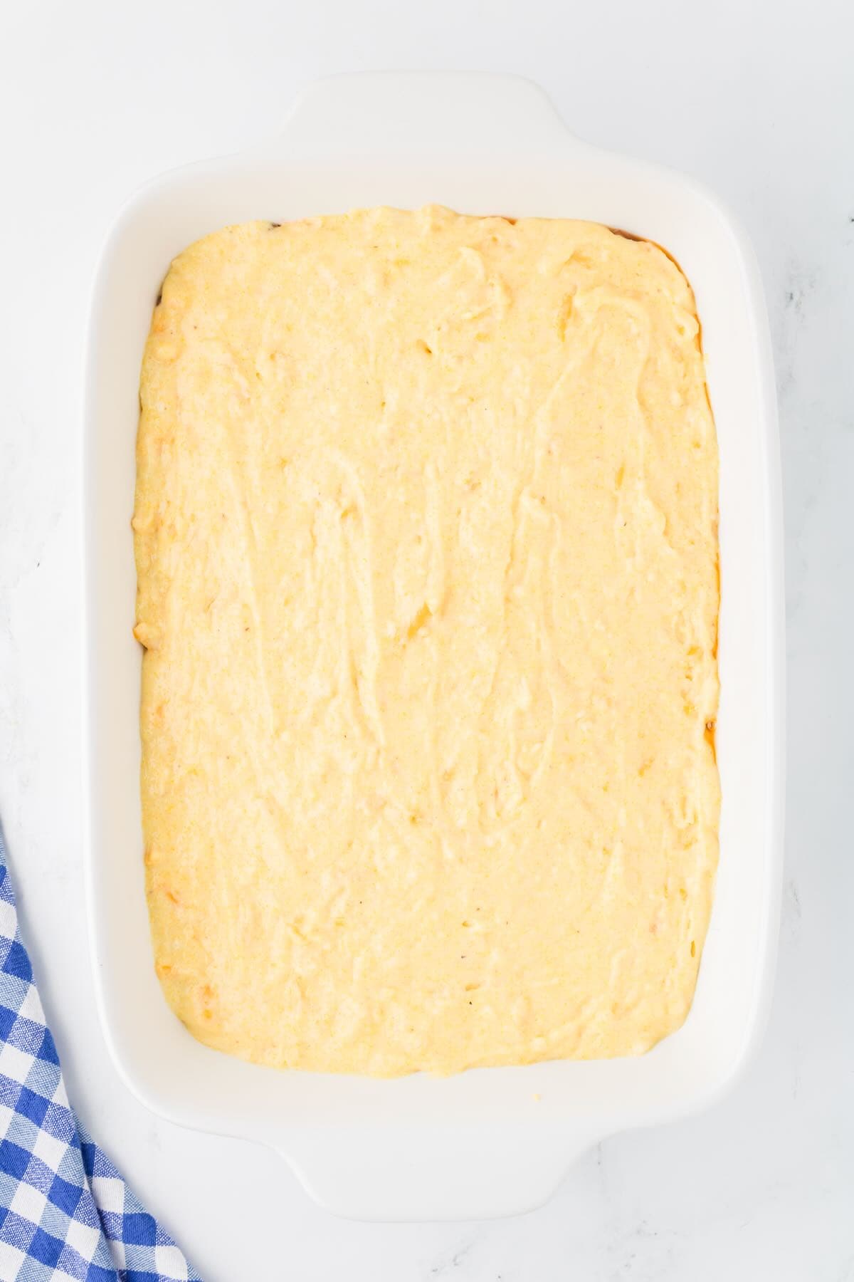 Cornbread batter spread out evenly over a beef filling in a white baking dish. 