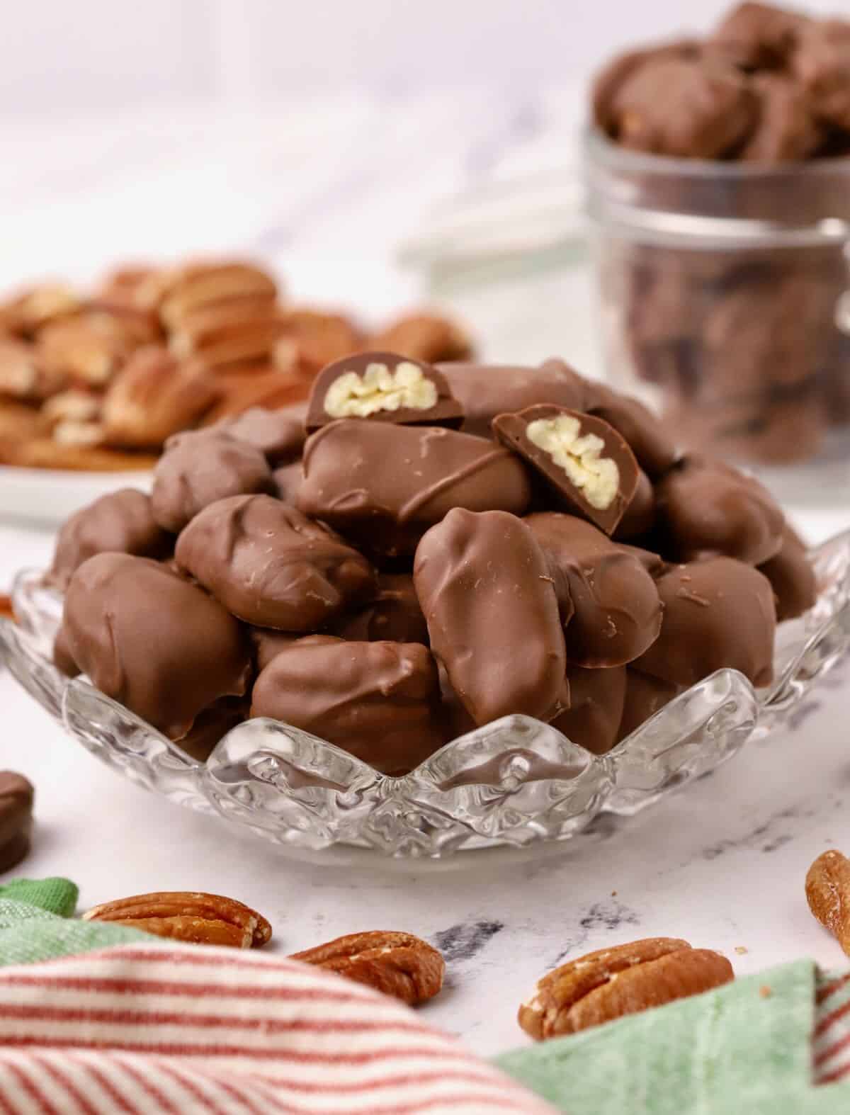 A small glass dish full of chocolate covered pecans and a dish of roasted pecans in the rear. 