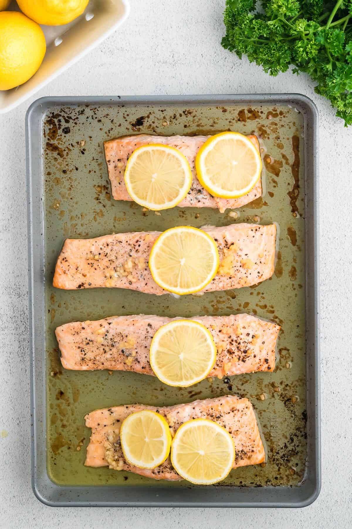 Four baked salmon fillets on a baking sheet topped with slices of lemon. 
