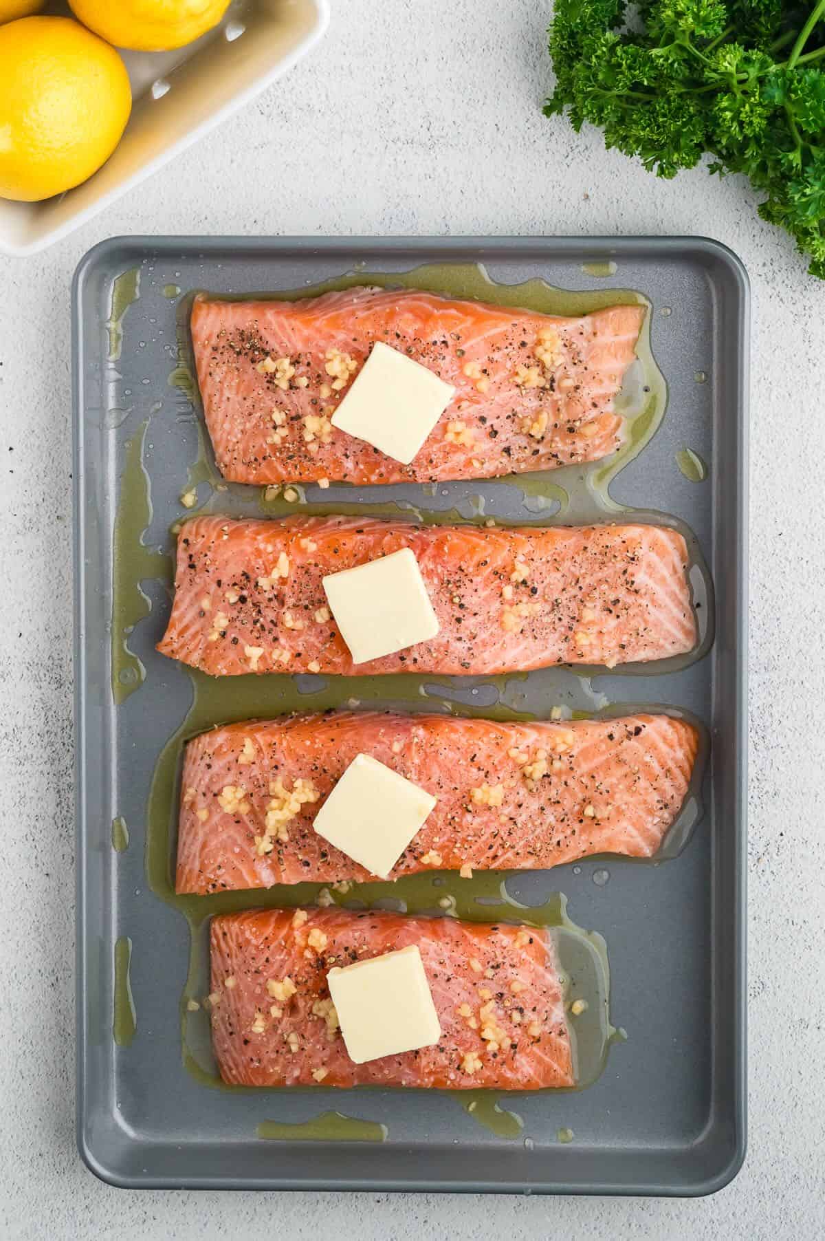 Four salmon fillets on a baking sheet, each topped with a pat of butter. 