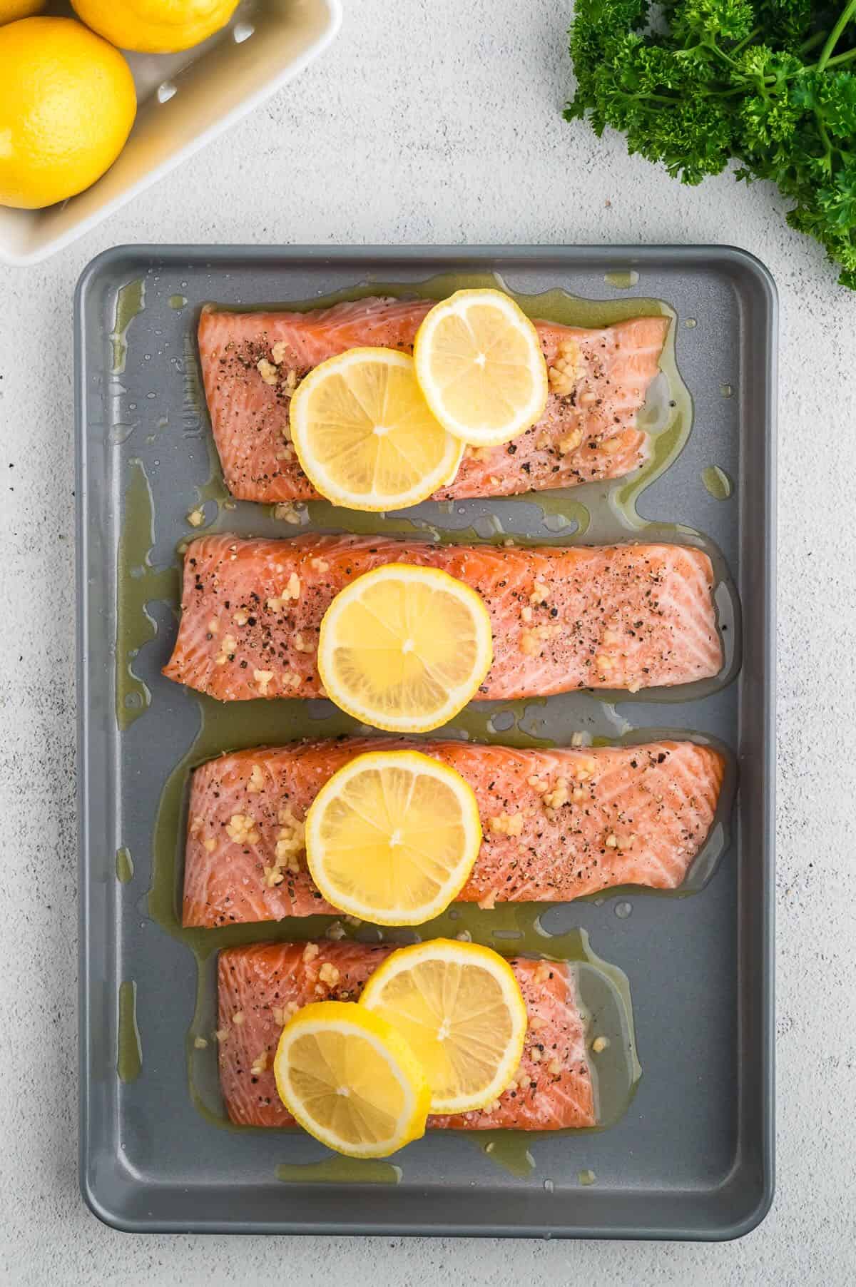 Four salmon fillets on a baking sheet topped with slices of lemon. 