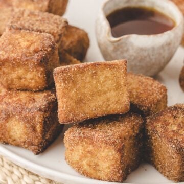 French toast bites stacked on a plate with a pitcher of syrup.