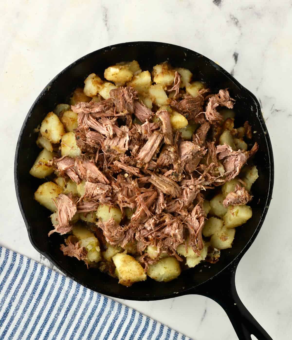 Cooked cubed potatoes in a cast iron skillet topped with shredded pot roast. 
