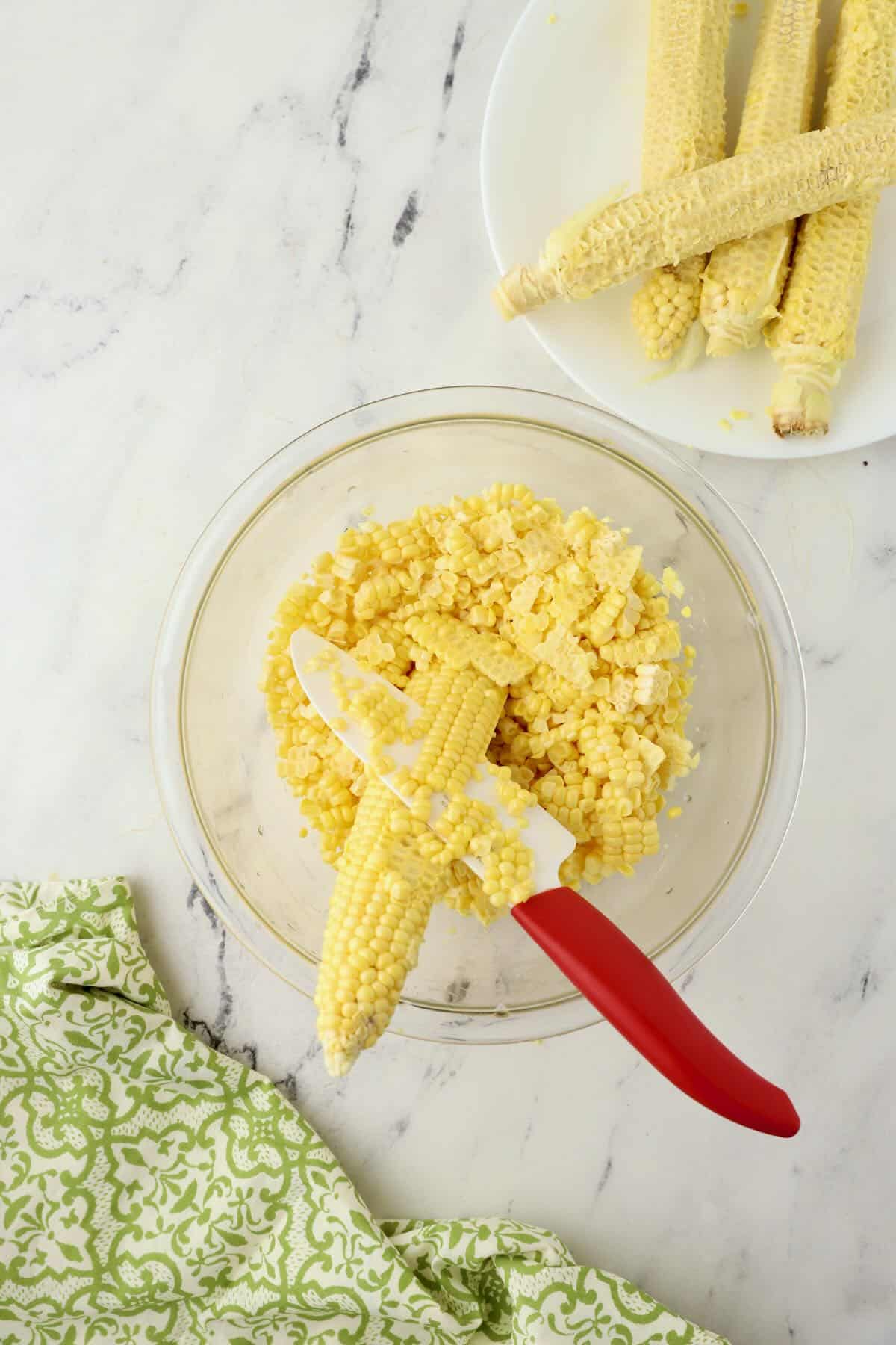 Cutting kernels off of a corn cob into a clear glass bowl using a knife.