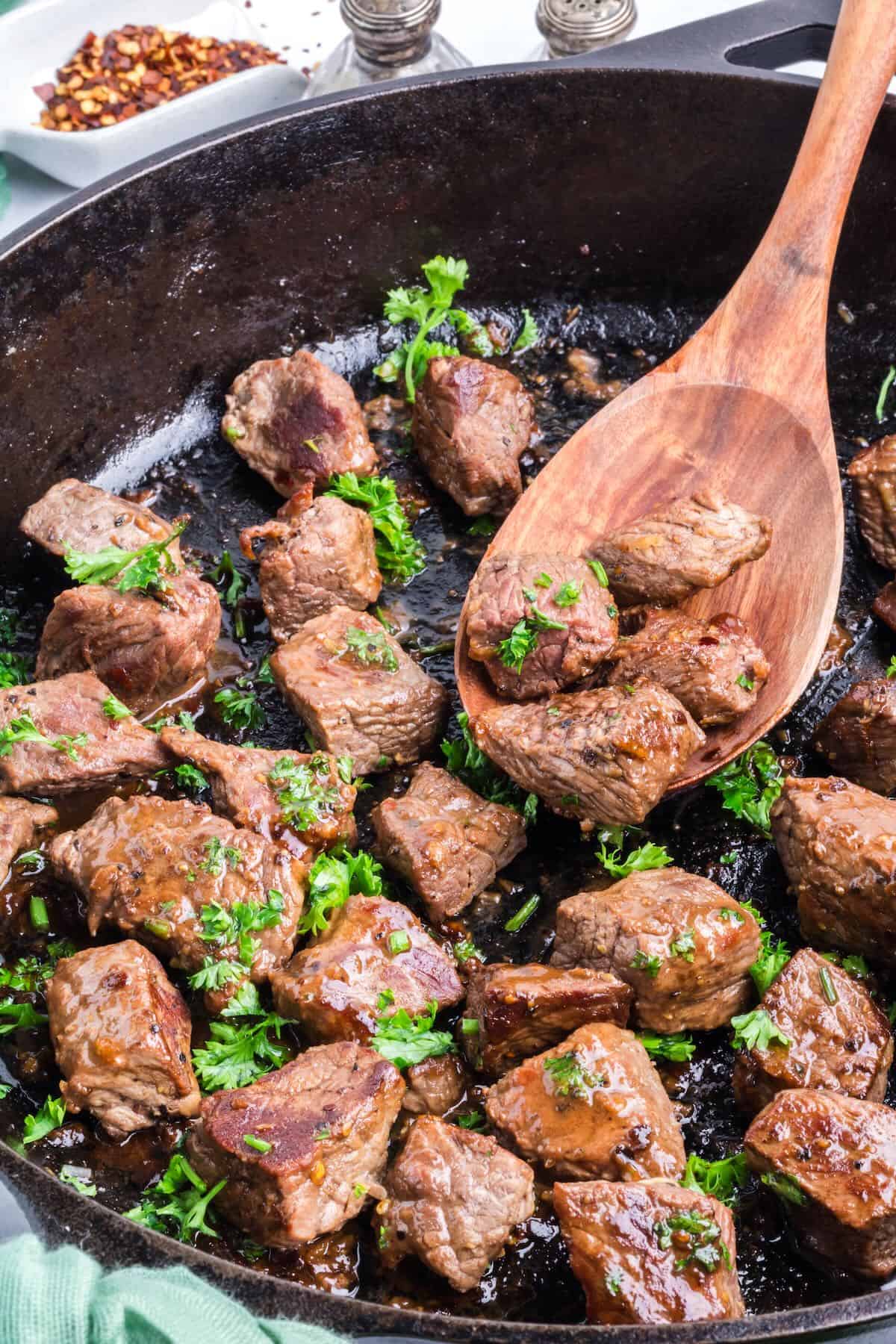 Steak bites in a cast iron skillet with a few in a wooden spoon. 