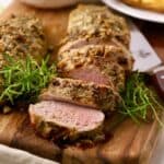 Sliced herb crusted pork tenderloin on a cutting board, garnished with sprigs of rosemary.