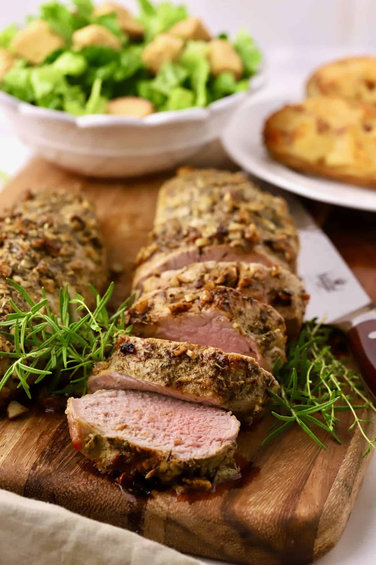 Herb crusted pork tenderloin sliced on a cutting board and garnished with sprigs of rosemary.