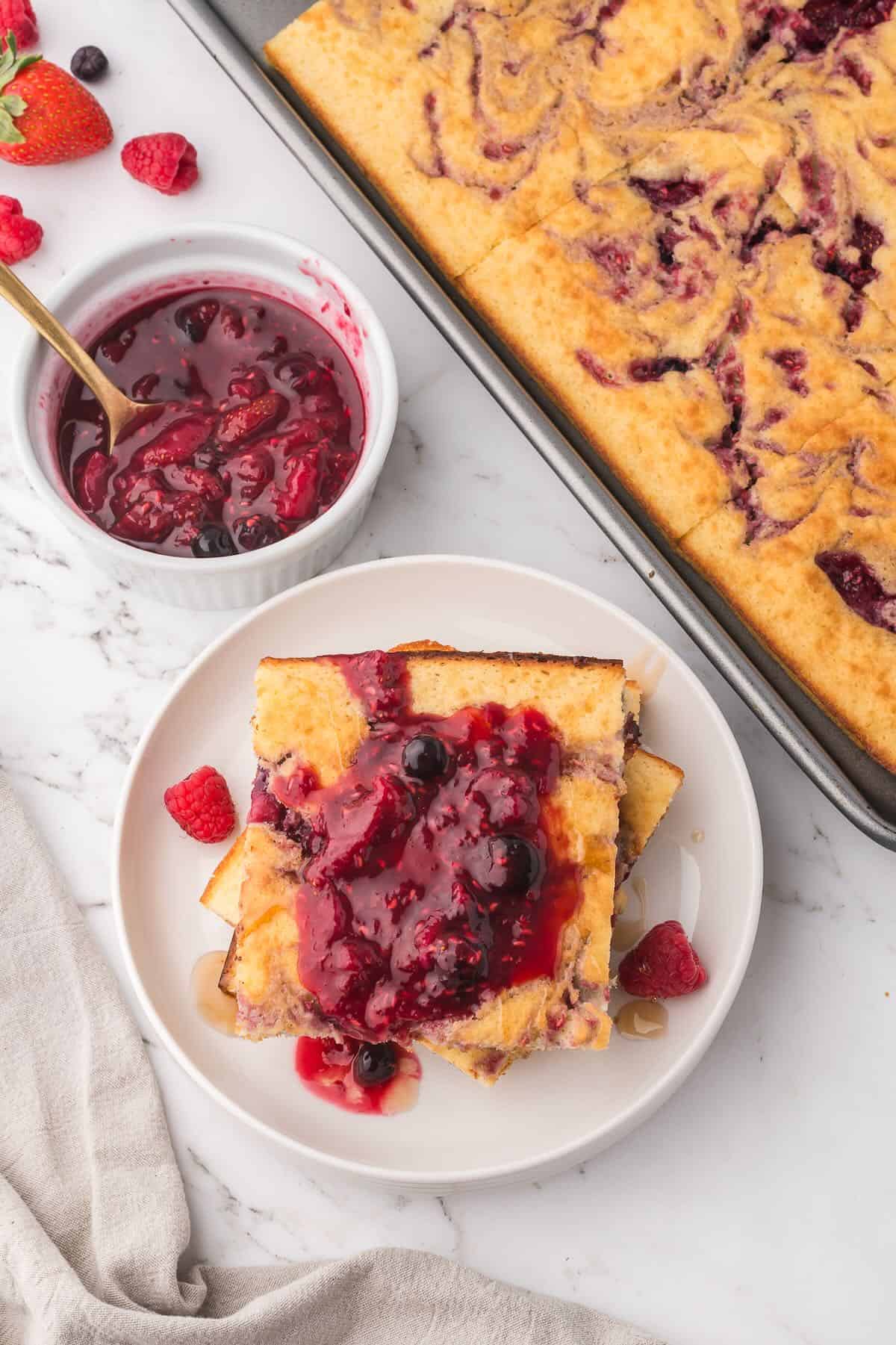 A stack of pancakes topped with berry compote, next to a sheet pan with pancakes/.