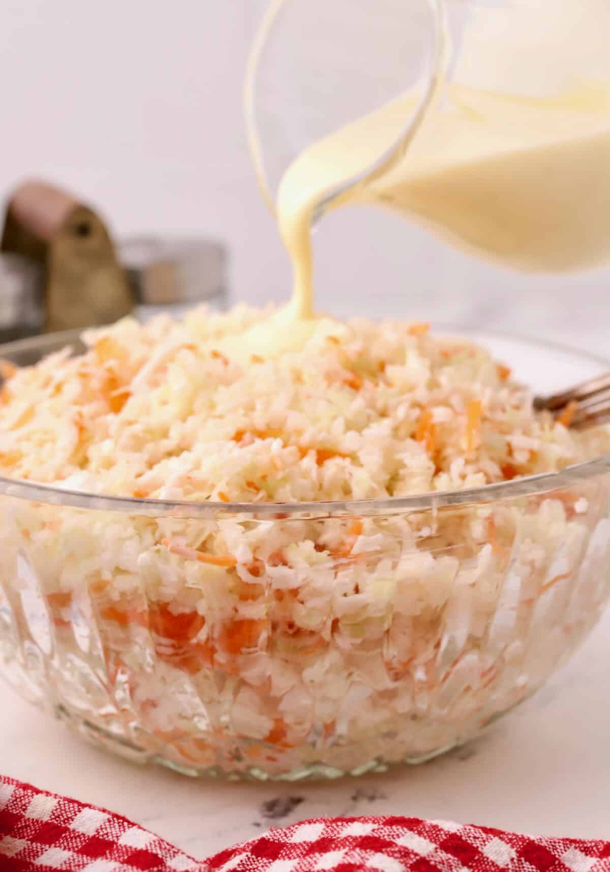 Pouring coleslaw dressing from a small pitcher into a large bowl of coleslaw. 