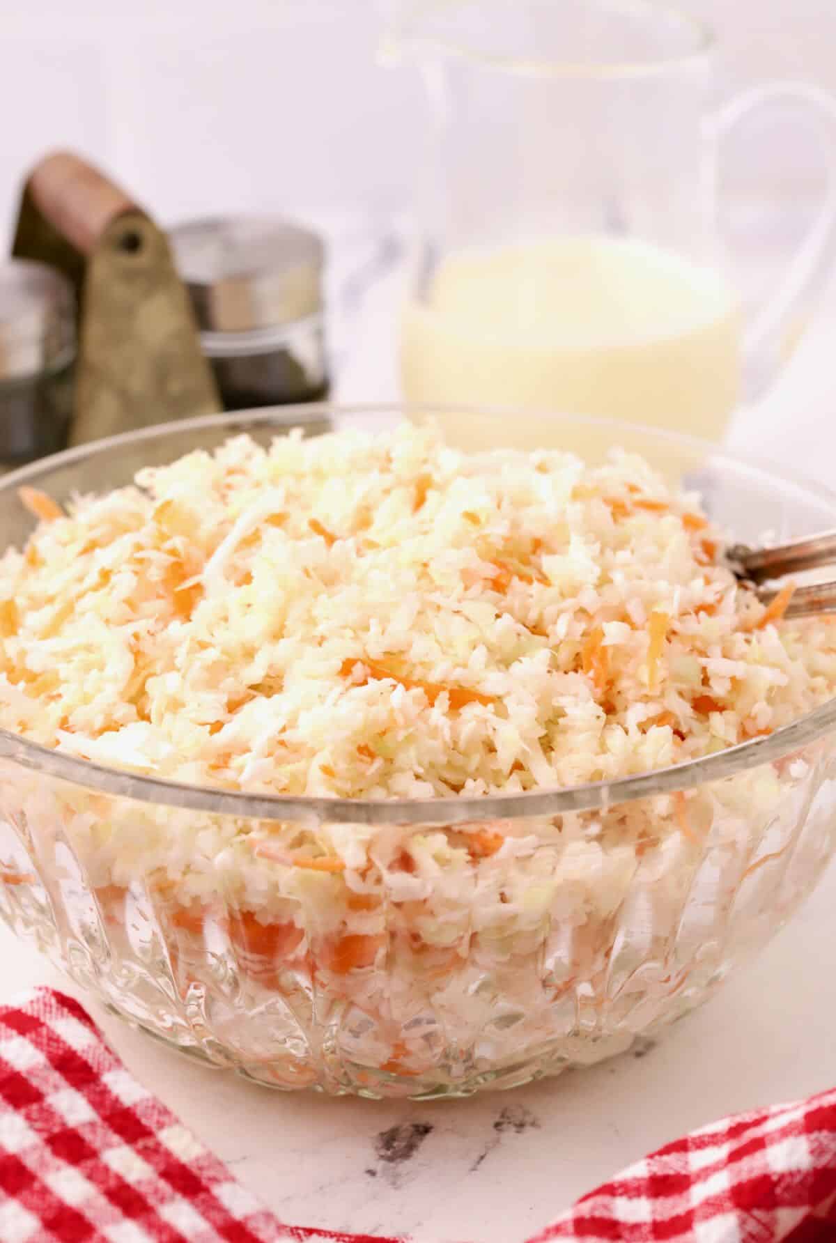A large glass bowl filled with coleslaw with a pitcher of dressing behind it. 