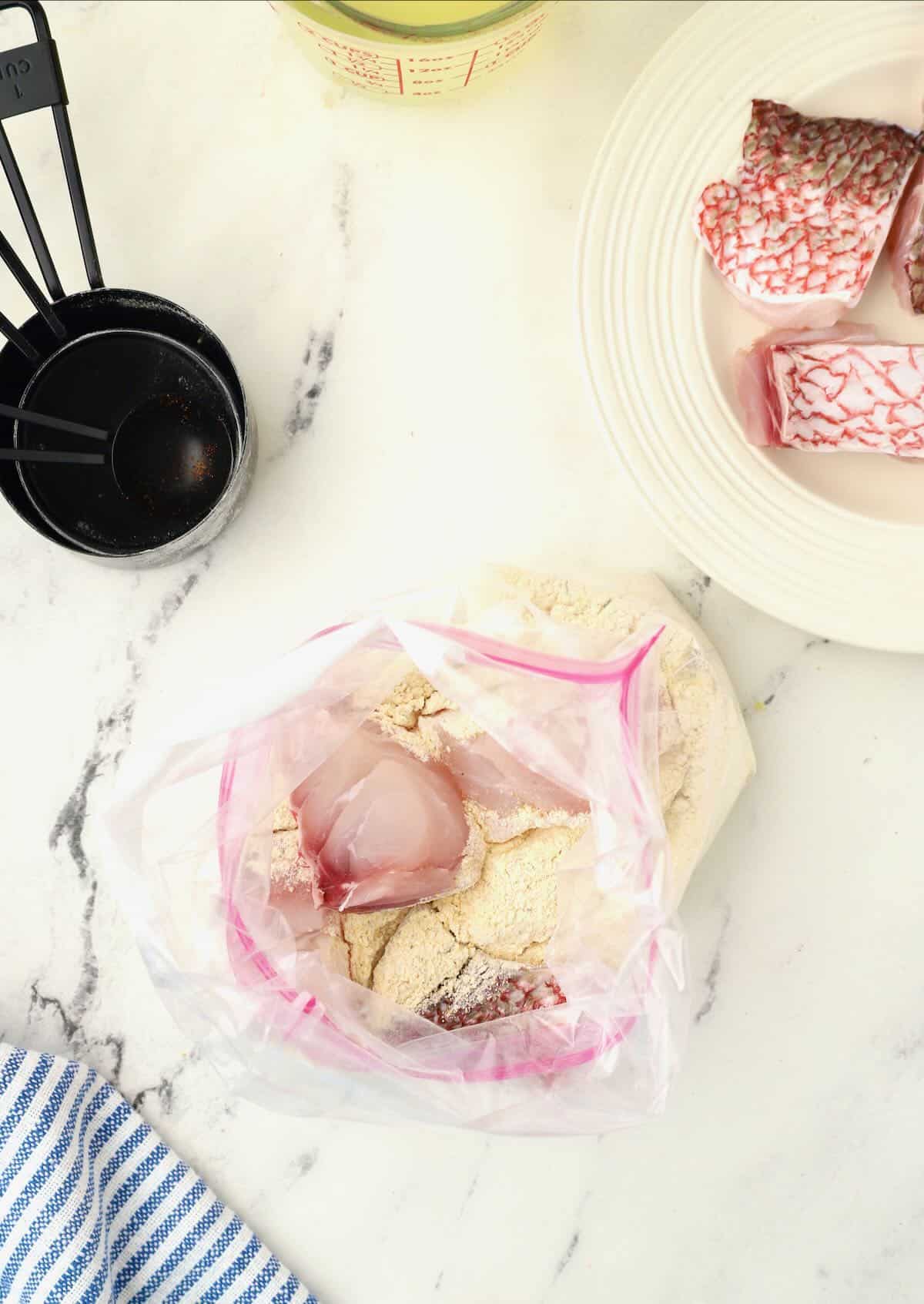 A food storage bag full of cornmeal, flour, and Creole seasonings with pieces of fish in it. 