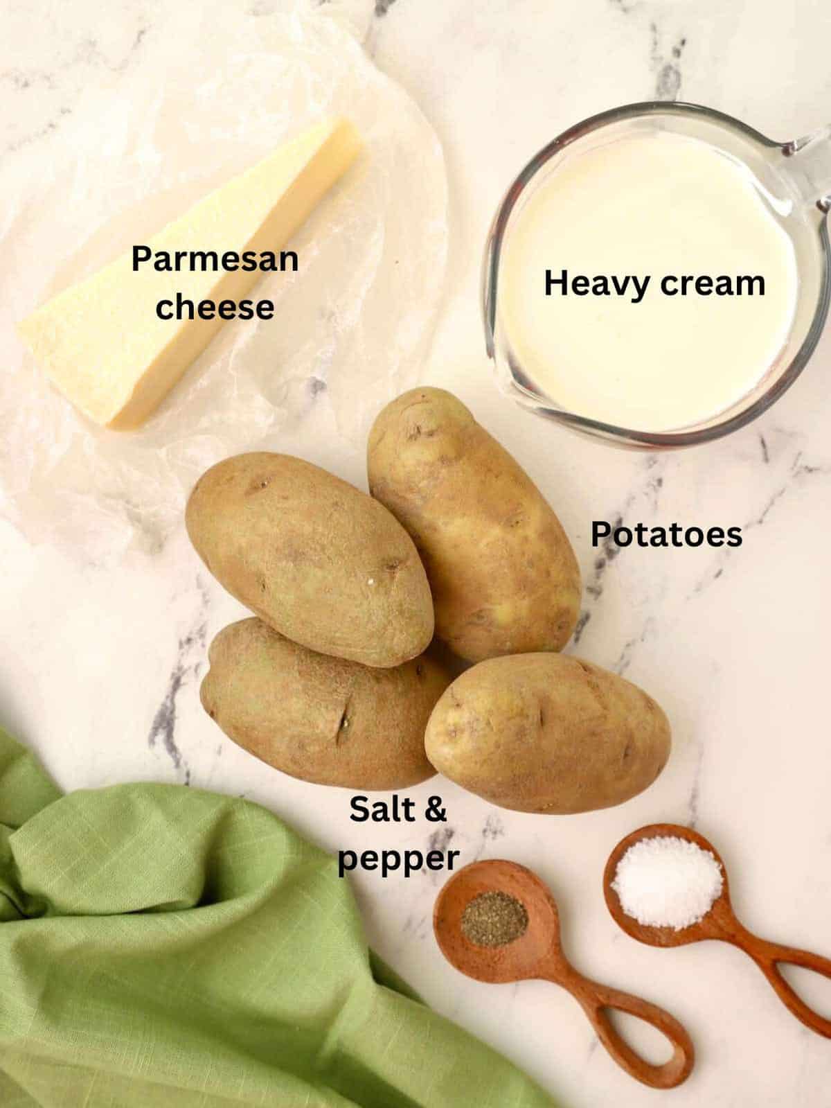Four potatoes, a measuring cup of heavy cream, and a block of parmesan cheese on a counter. 