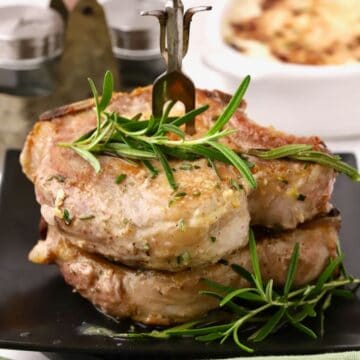Two pork chops stacked with a sprig of rosemary on top and a fork sticking out.