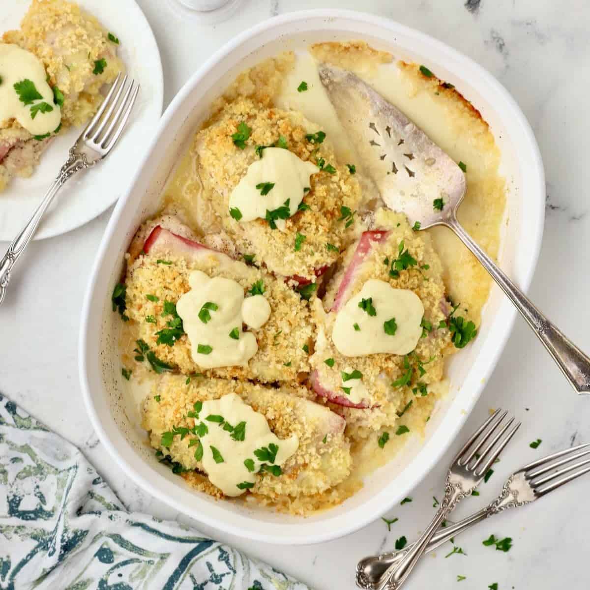 A white baking dish full of baked chicken cordon bleu casserole topped with sauce. 
