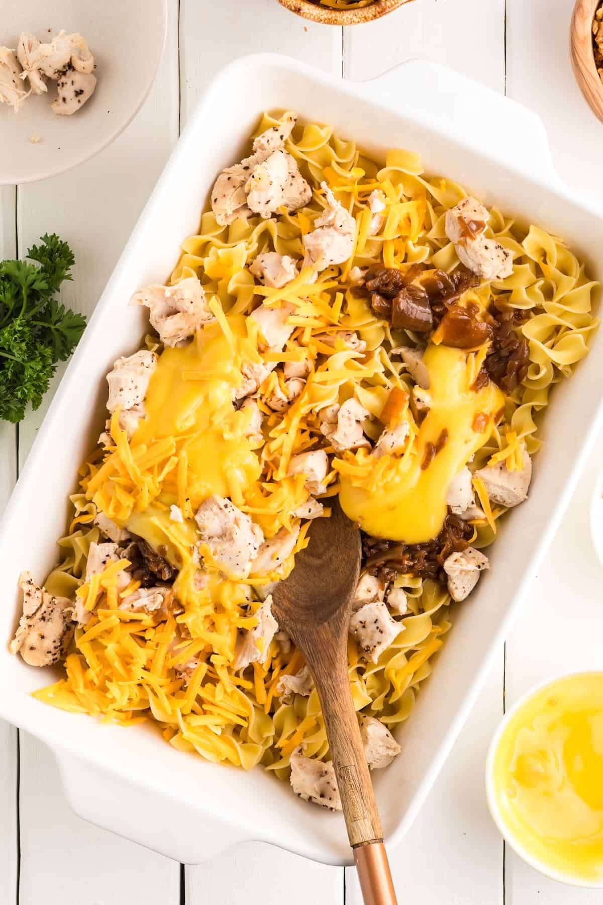 All of the ingredients for French Onion Soup Chicken Casserole in a white baking dish being mixed with a wooden spoon.