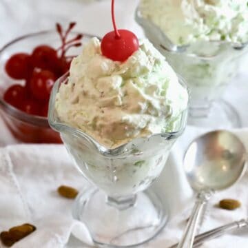Pistachio pudding dessert in a tall glass dessert dish, topped with a cherry.
