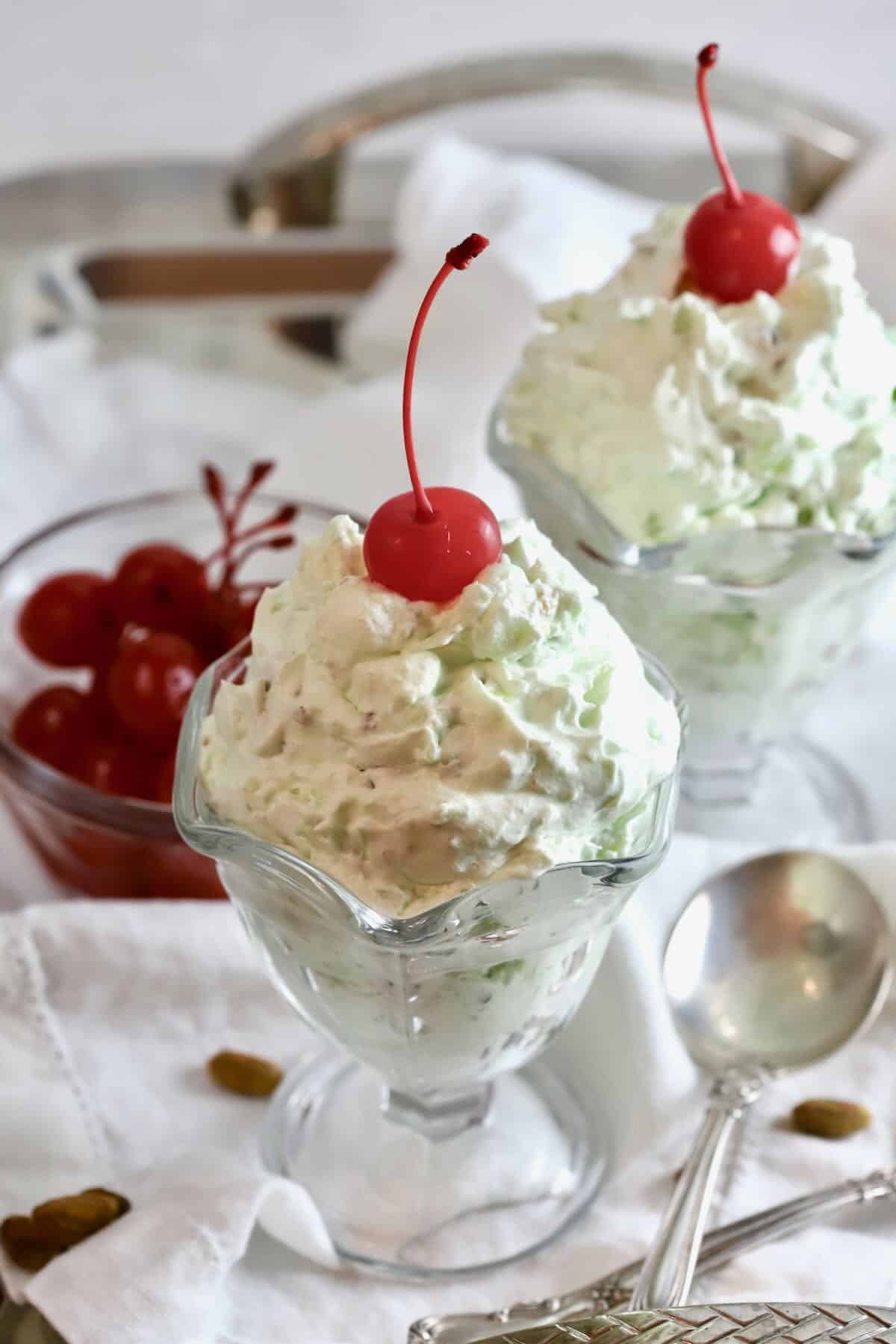 Two pistachio pudding desserts in a tall glass dessert dish, topped with a cherry. 