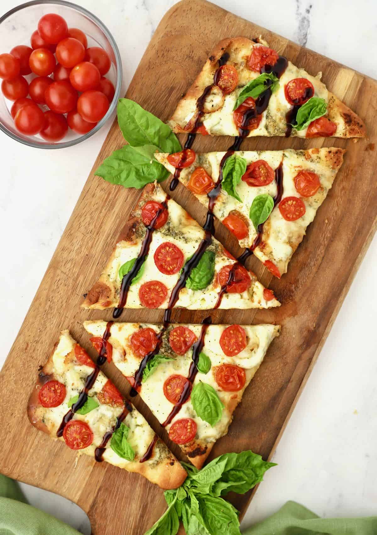 A cutting board with sliced caprese flatbread topped with cherry tomatoes, balsamic glaze and fresh basil leaves. 