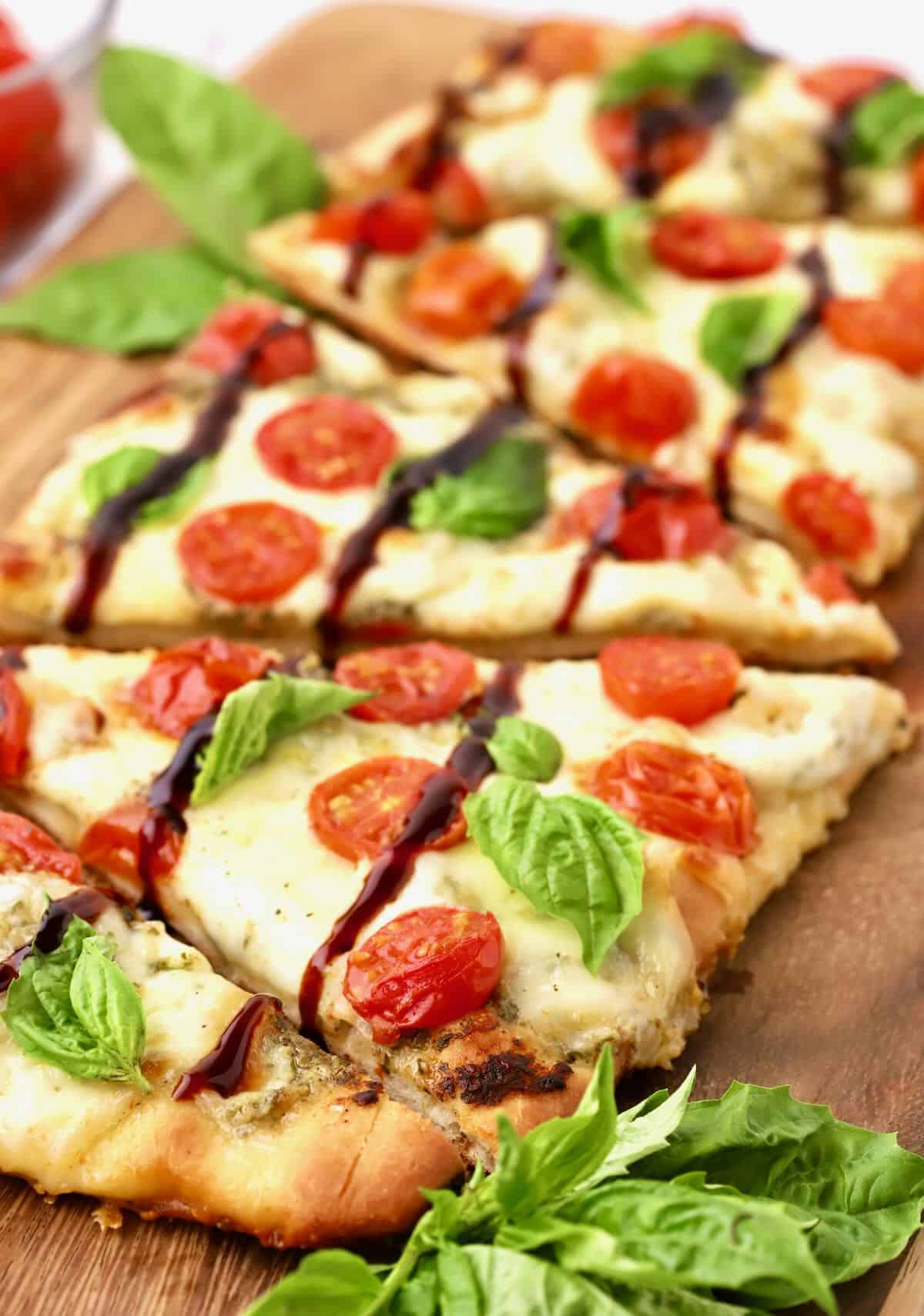 Caprese flatbread on a cutting board cut into slices with balsamic glaze and basil leaves. 