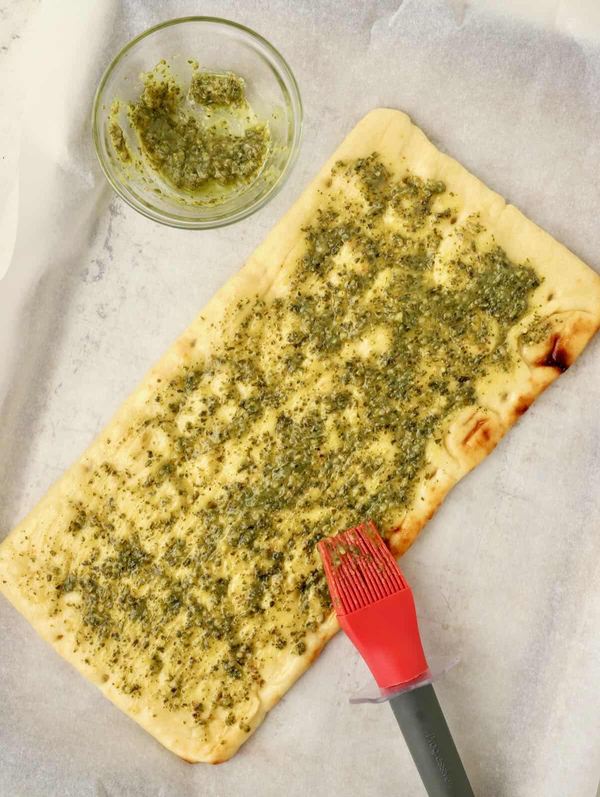 A flatbread with basil pesto being brushed on the top. 