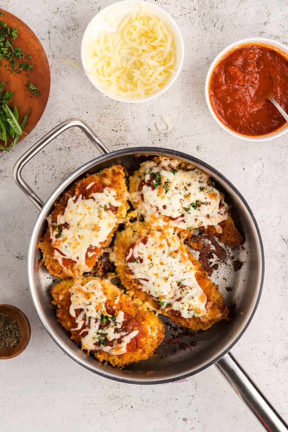 Four fried chicken breasts in a skillet topped with marinara sauce and melted cheese. 