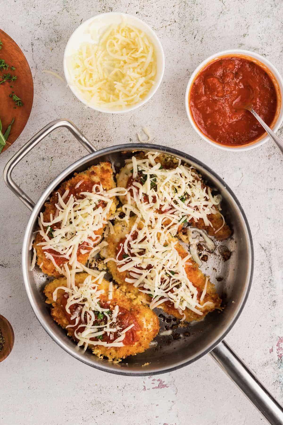Four fried chicken breasts in a skillet topped with marinara sauce and cheese. 