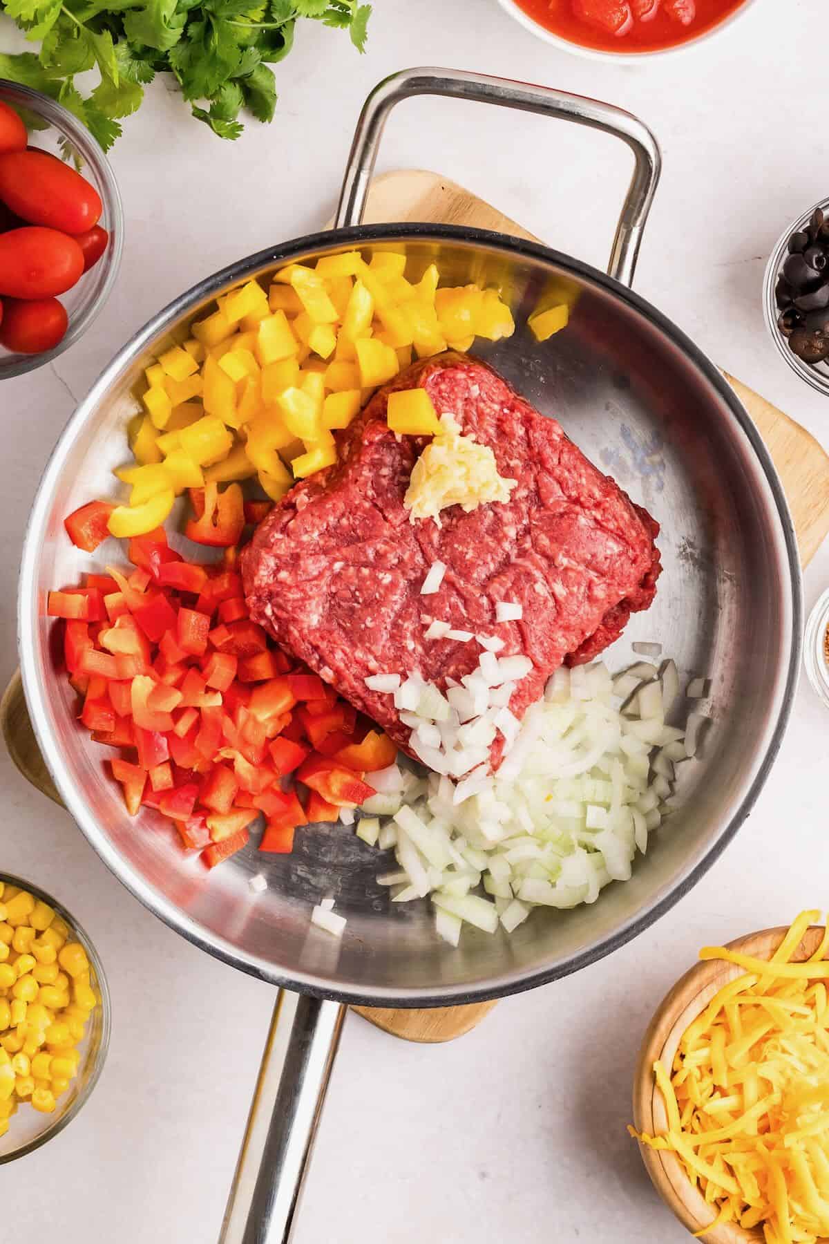 A block of ground beef and chopped onions and bell peppers in a skillet. 