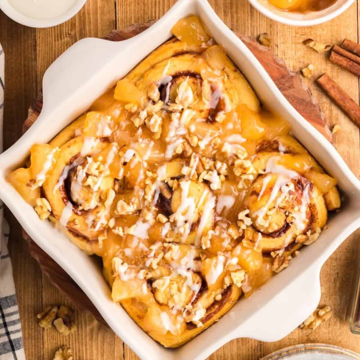 Apple Pie Filled Cinnamon Rolls in a white square baking dish topped with chopped apples, nuts, and icing.