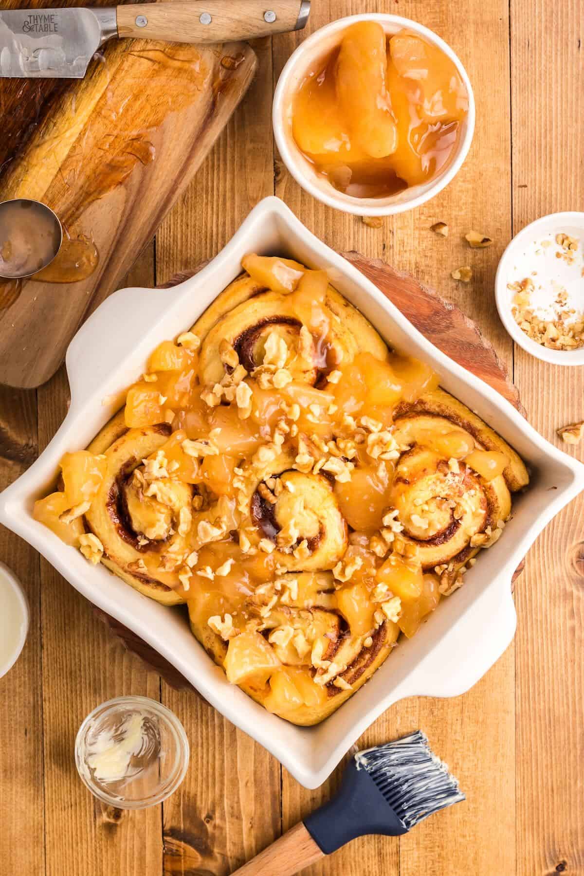 Baked cinnamon rolls in a baking dish topped with chopped apples and nuts. 
