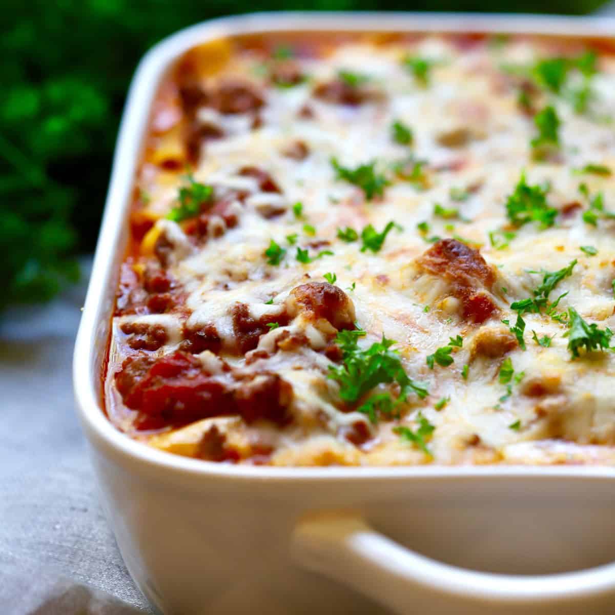 Baked ziti in a white baking dish topped with melted cheese and garnished with parsley. 