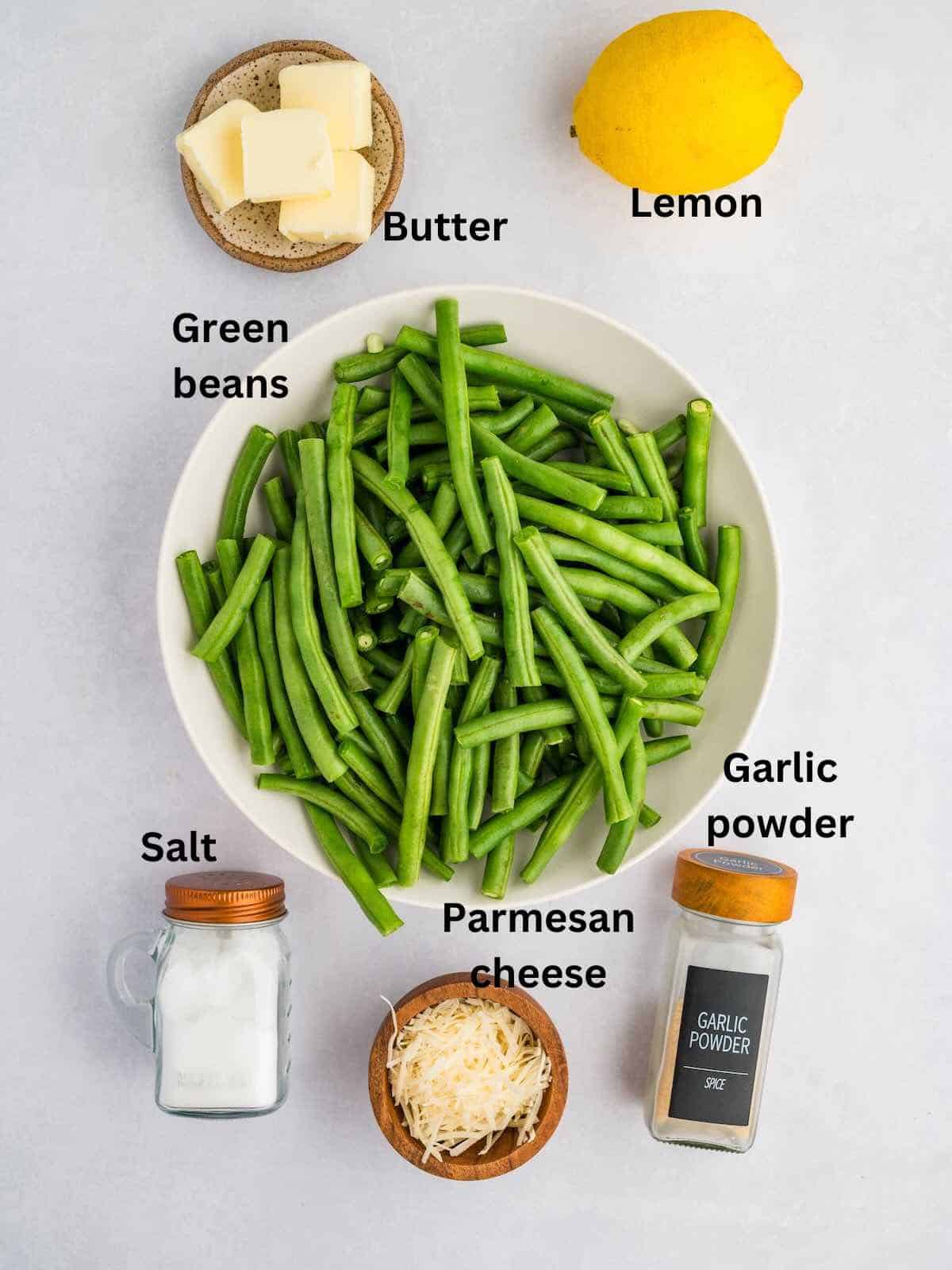A bowl of green beans, jars of salt and garlic salt, pats of butter, a lemon and pats of butter. 