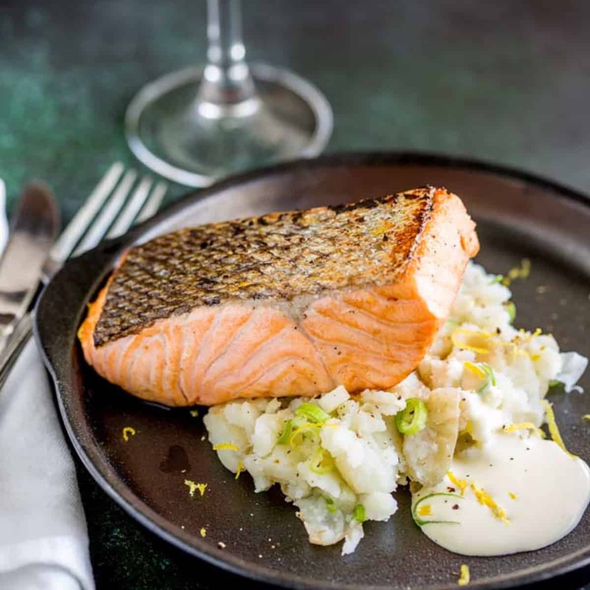 A piece of pan-seared salmon on top of mashed potatoes on a black plate. 