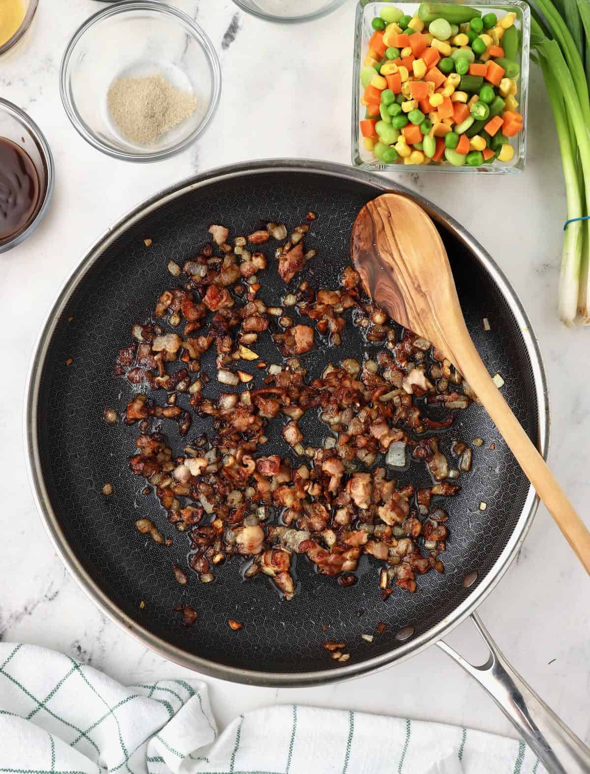 Chopped up bacon and onions cooking in a large skillet. 