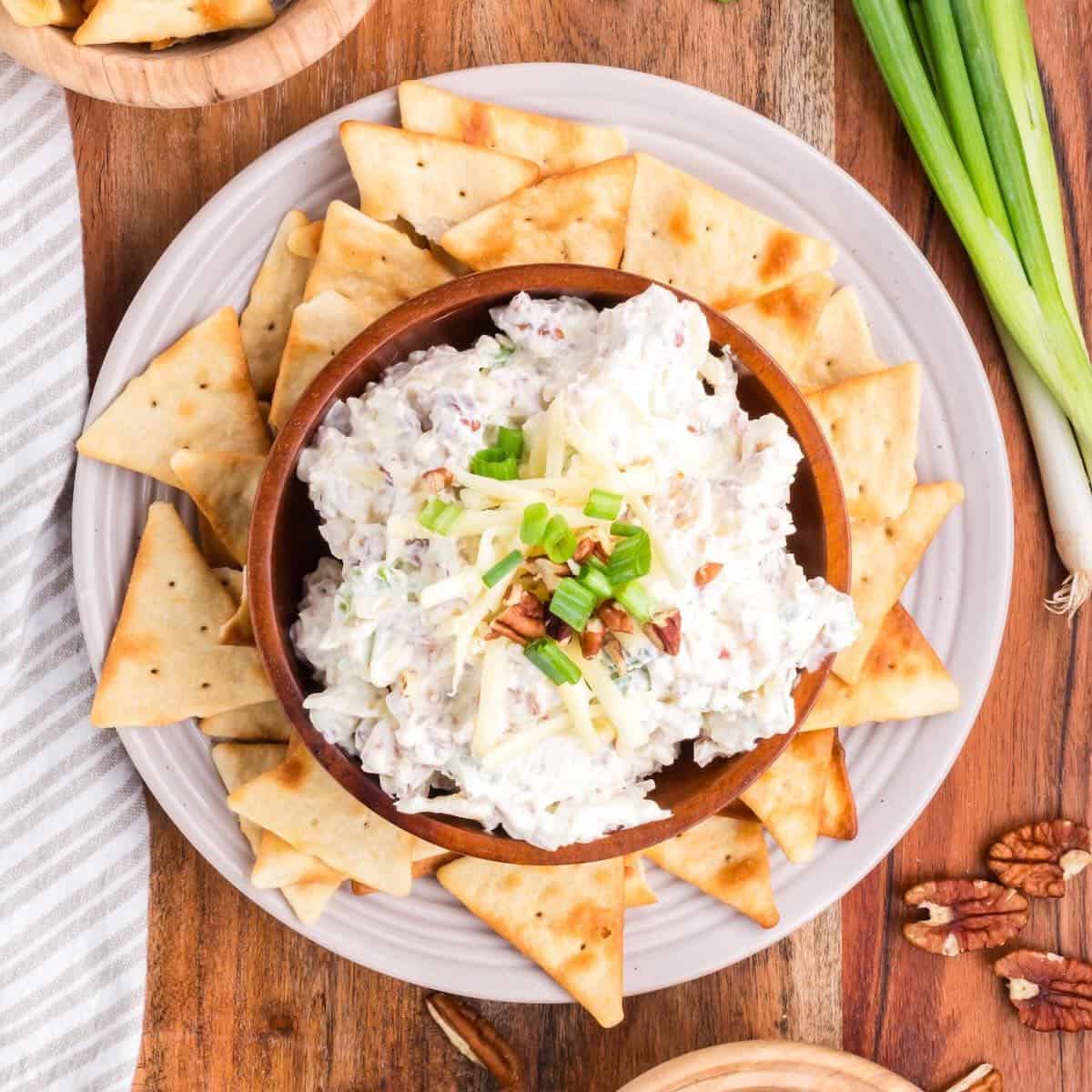 Bacon cream cheese dip on a plate with crackers and garnished with pecans and chopped-up scallions.
