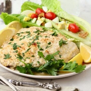 A baked grouper fillet topped with sour cream and parmesan cheese and garnished with parsley and sliced lemons on a plate.