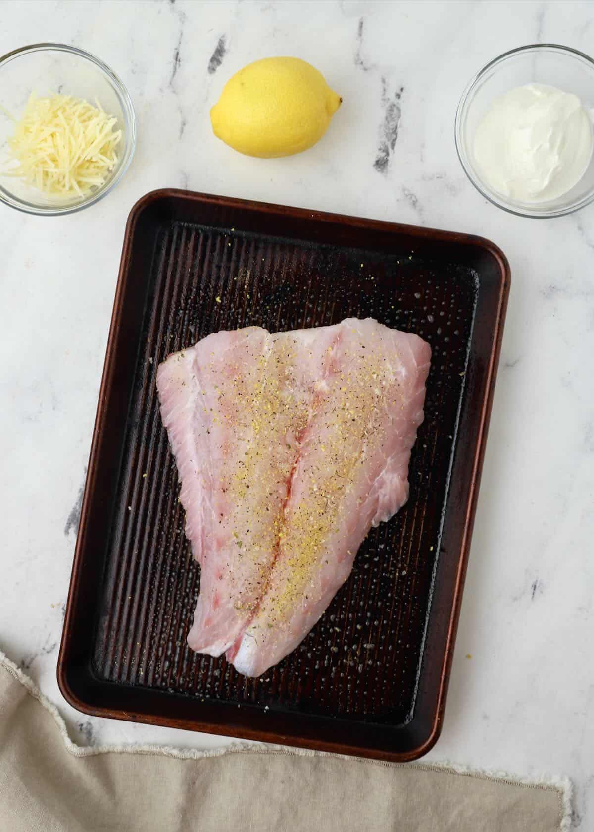 A large grouper fillet seasoned with salt and lemon pepper on a baking sheet. 