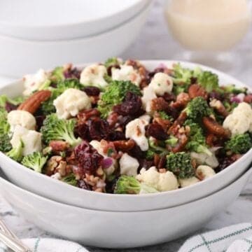 A white serving bowl with broccoli, cauliflower, and cranberry salad topped with pecans.
