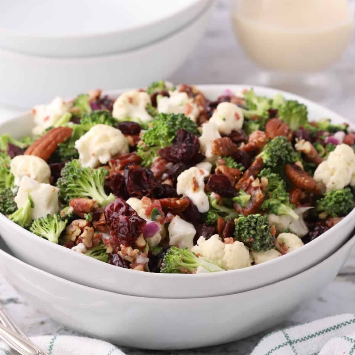 A white serving bowl with broccoli, cauliflower, and cranberry salad topped with pecans.