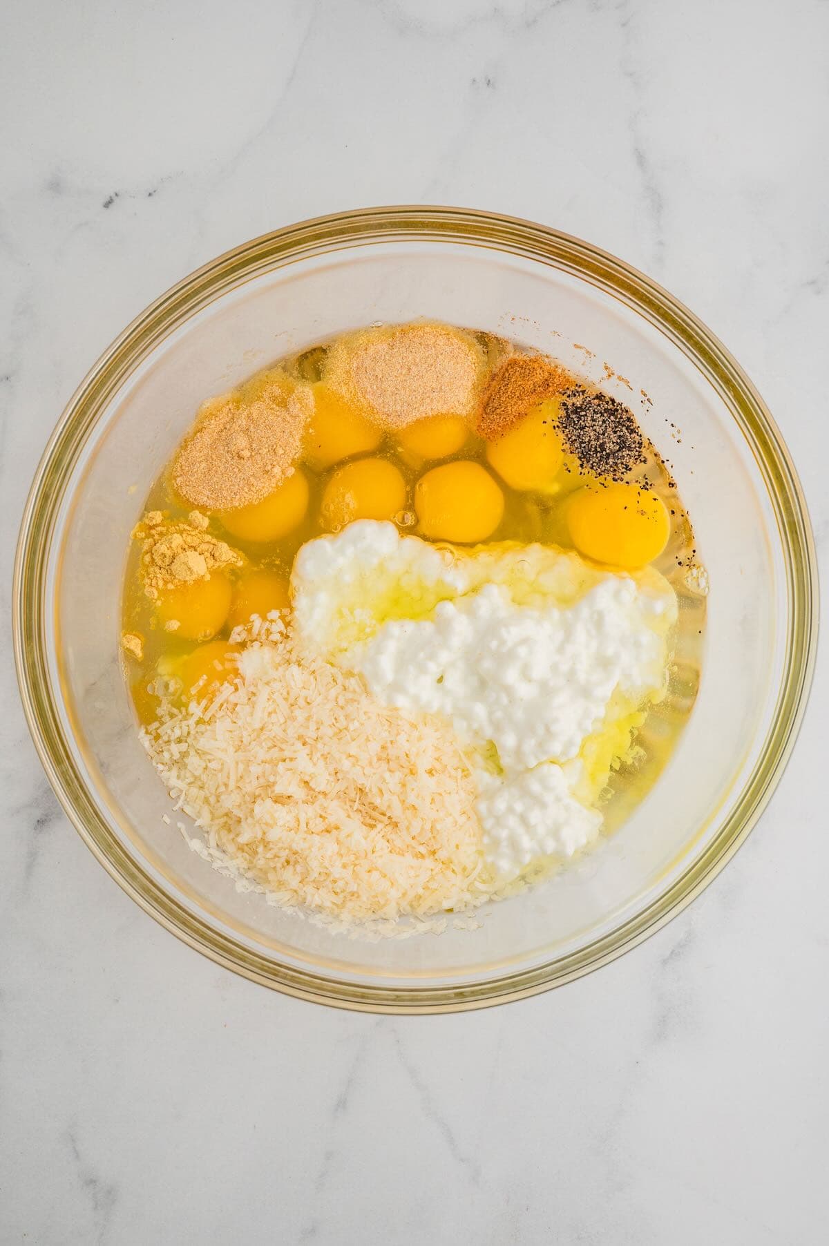 A dozen eggs, cottage cheese and several different seasonings in a clear glass bowl. 