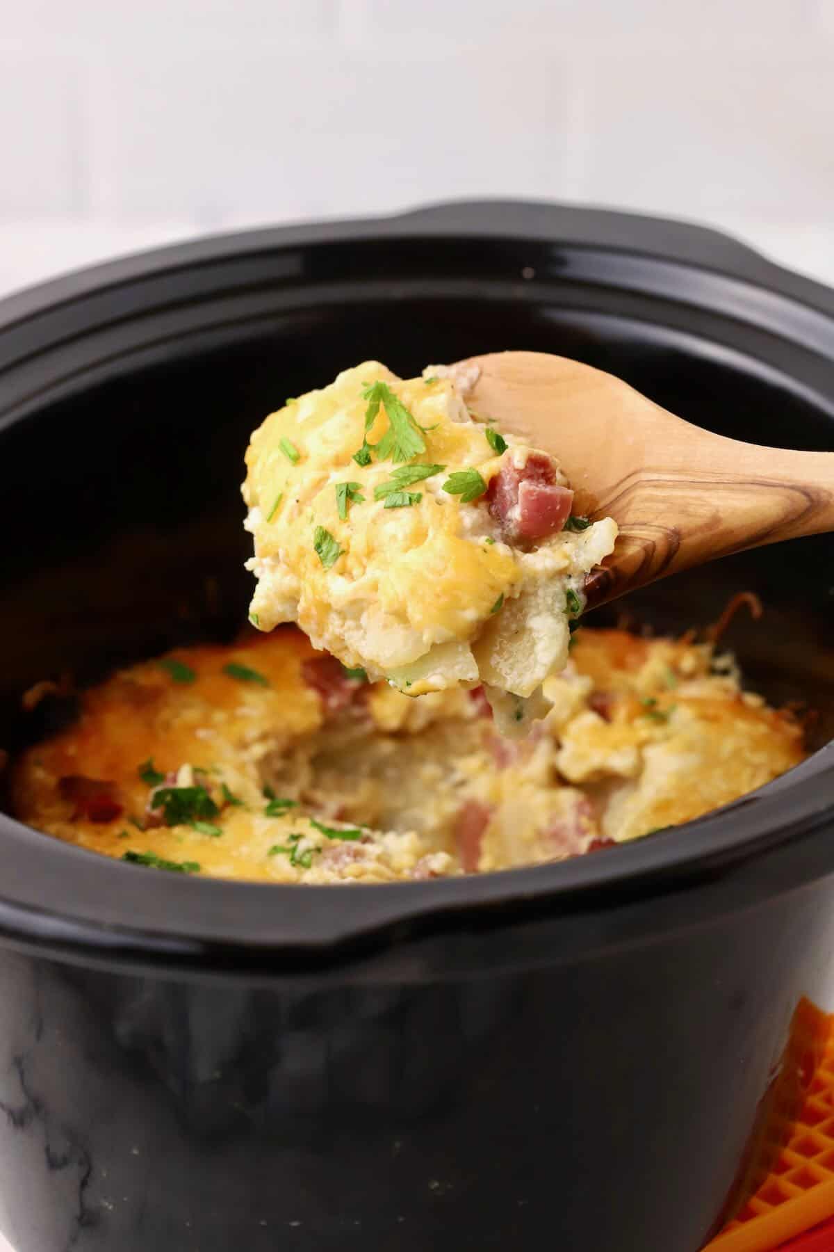 Ham and scalloped potatoes cooked in a crock pot, showing a wooden spoon with a spoonful. 