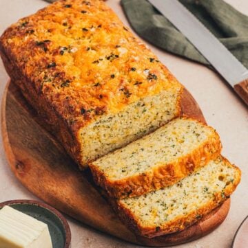 A loaf of savory cheese bread with herbs on a cutting board with slices cut.
