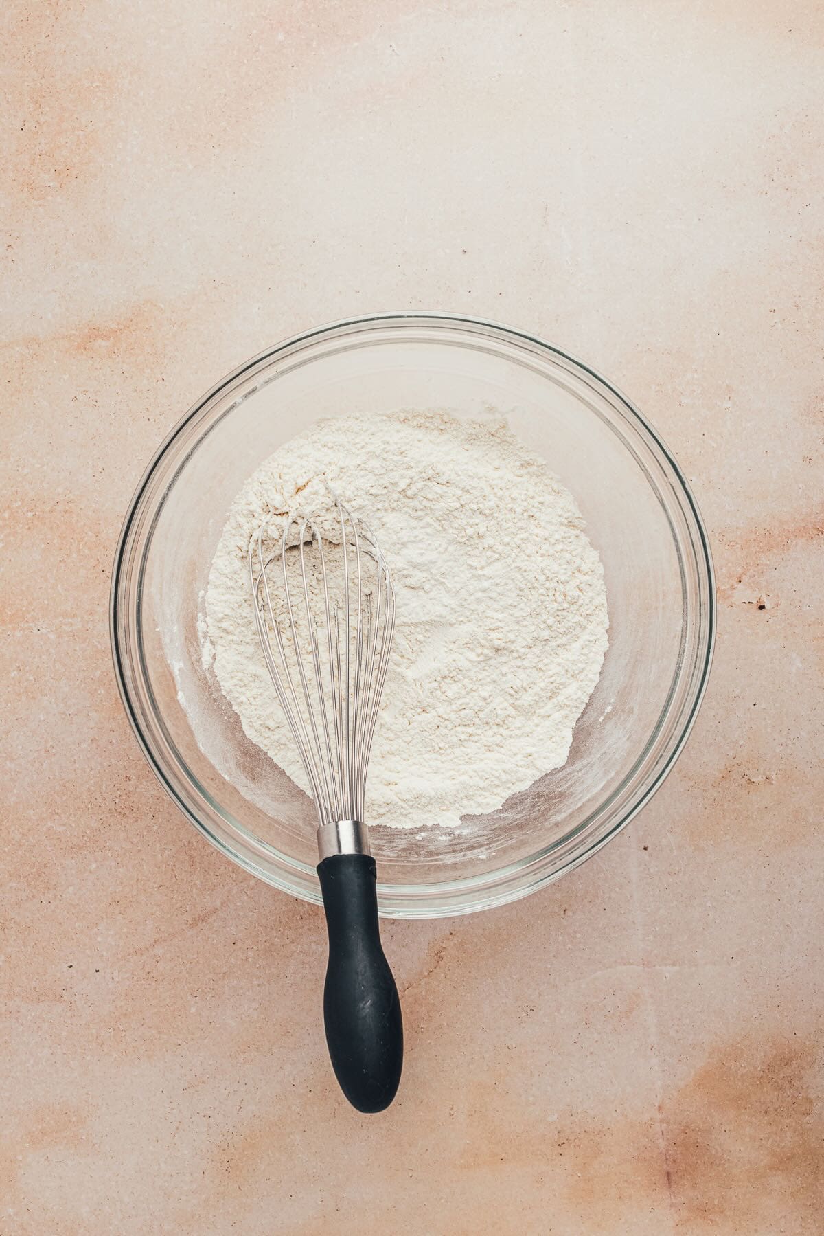 Flour and other dry ingredients in a clear glass bowl with a whisk. 