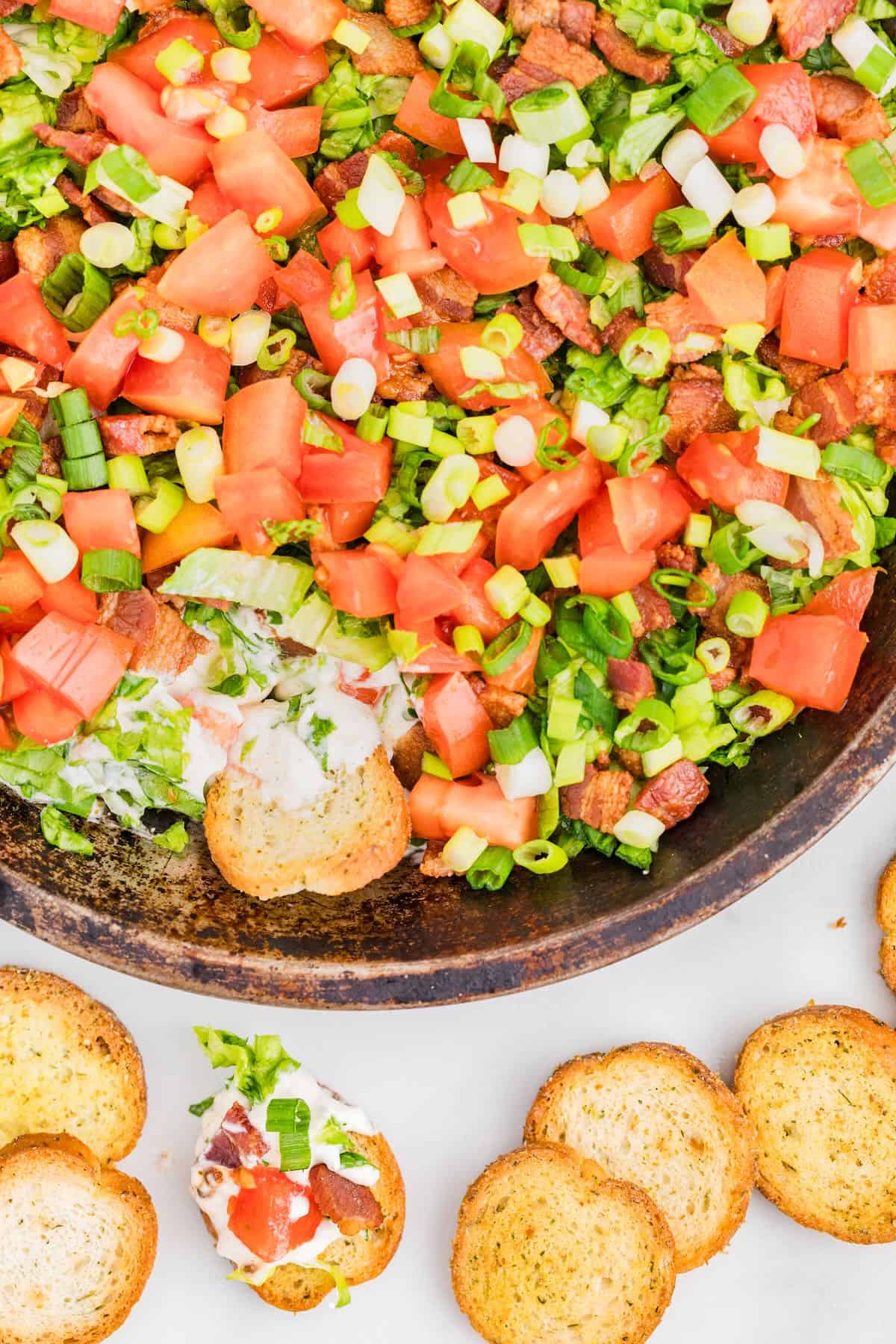 BLT Dip in a wooden bowl topped with bacon, lettuce, and tomatoes with toasted baguettes.