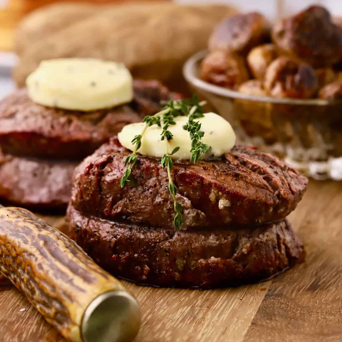 A grilled filet mignon topped with a pat of butter and thyme sprigs on a cutting board.
