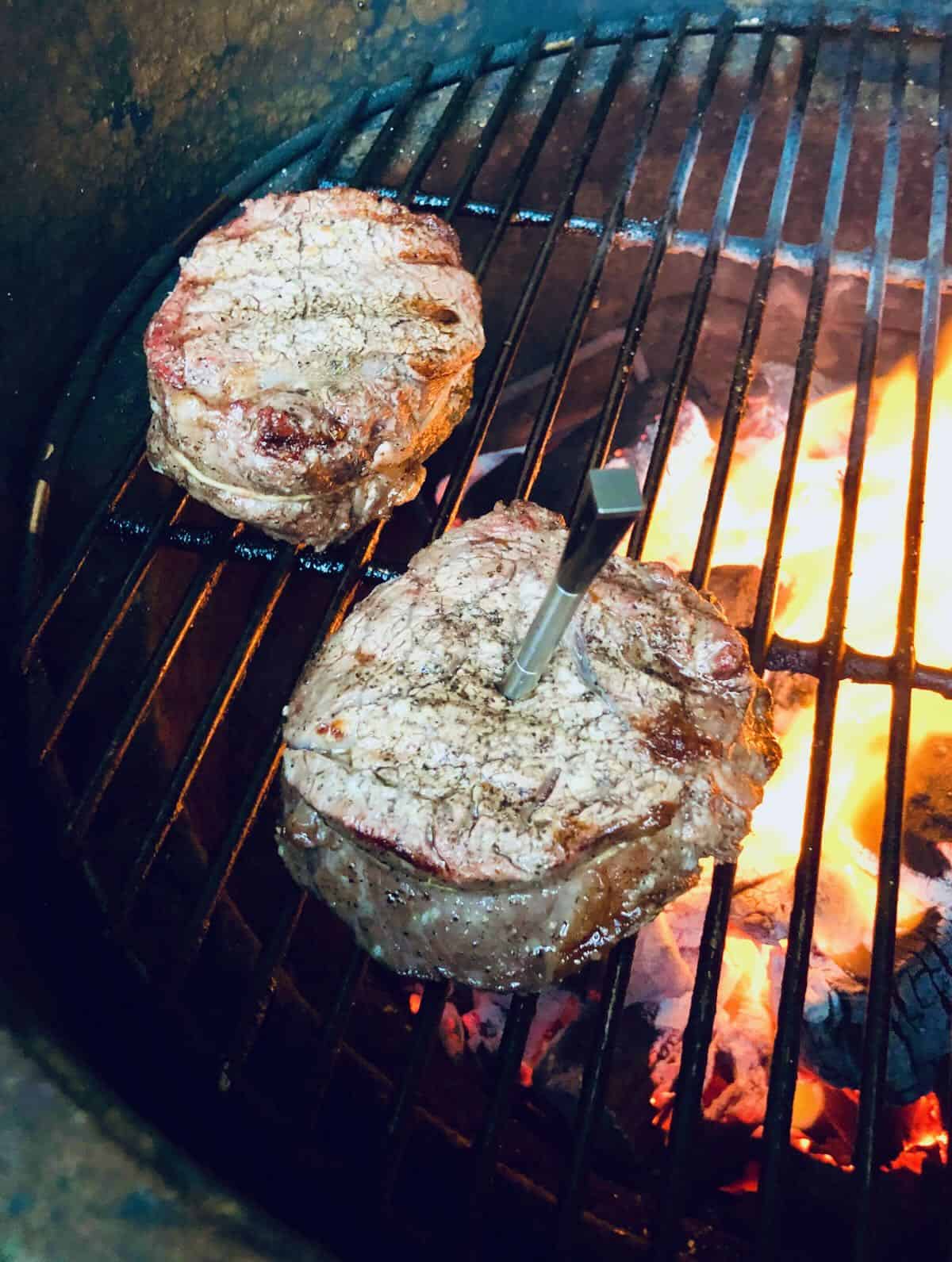 Two steaks cooking on a charcoal grill, one with a meat thermometer in the middle. 