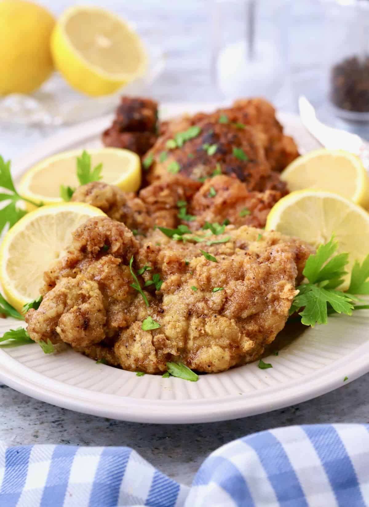 A white platter with fried lemon pepper chicken thighs garnished with sliced lemons. 